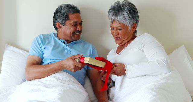 A middle-aged Latino couple shares a joyful moment as the man presents a gift to the woman, with copy space. Their smiles and the cozy bedroom setting suggest a celebration of a special occasion or an intimate gesture of affection.