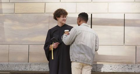 Graduate celebrating with friend outdoors by beige wall, holding mortarboard and coffee