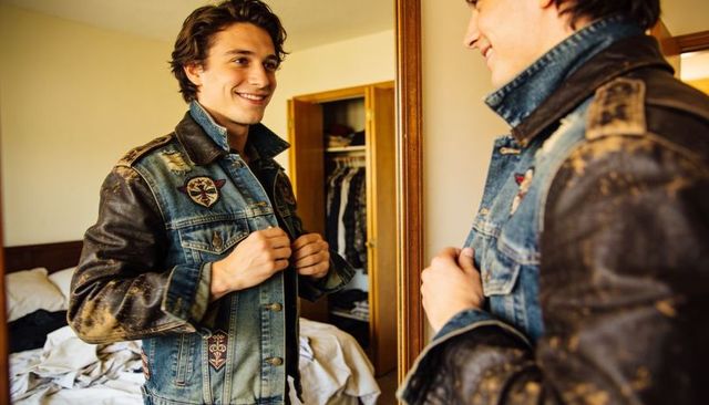 Young man fastening patched denim jacket while checking mirror in bedroom with rumpled bed