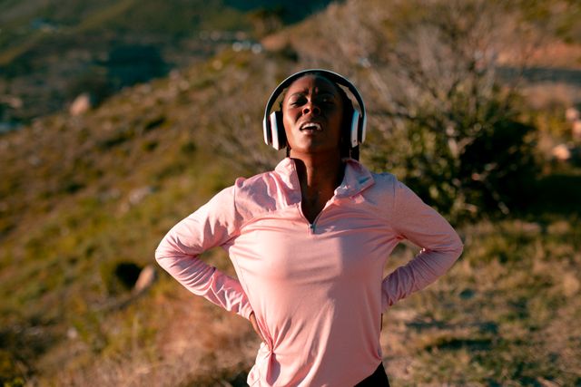 African American woman wearing headphones, engaging in outdoor exercise in a natural setting. Ideal for promoting healthy lifestyles, fitness routines, outdoor activities, and wellness programs. Suitable for use in advertisements, fitness blogs, and health-related publications.
