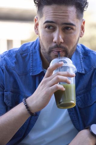 African american man sipping green smoothie on patio in denim jacket, thoughtful urban look