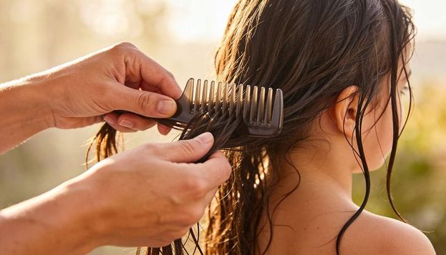Parent combing wet hair, detangling long strands with wide-tooth comb at sunlit poolside