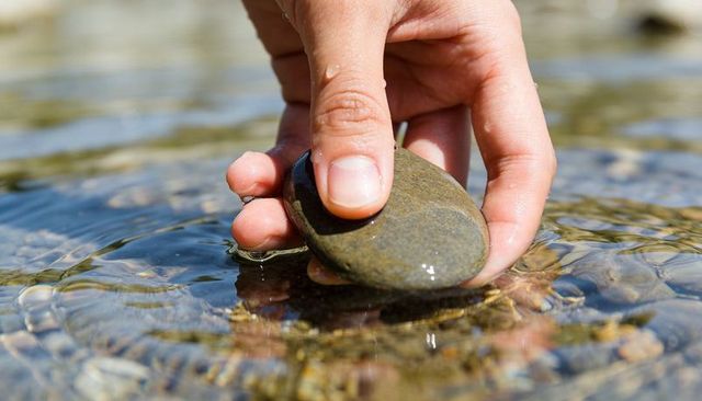 Hand Holding Smooth River Stone in Shallow Water with Ripples and Sunlight Reflection