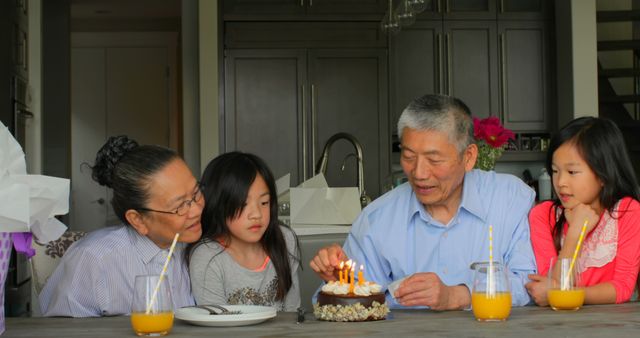 Multigenerational family enjoying a birthday celebration at home. Grandparents and children gathered around kitchen table with cake and lit candles, juice glasses in background. Perfect for promoting family values, special occasions, and home bonding moments.