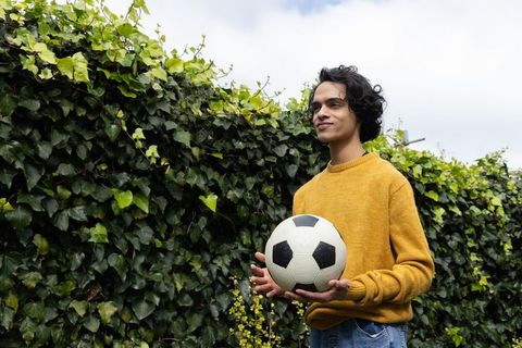 Asian teenager holding soccer ball by ivy hedge in mustard sweater