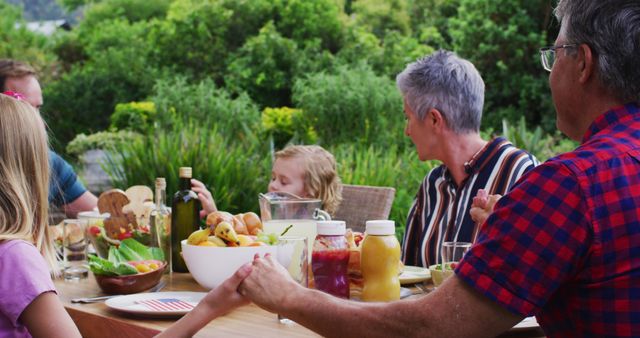 Smiling caucasian family holding hands saying grace before celebration meal together in garden. three generation family celebrating independence day eating outdoors together.