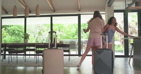 Laughing friends arriving at modern rental pulling suitcases through bright open living area