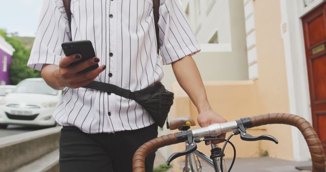 Biracial young professional holding smartphone, standing with bike. Wearing striped shirt, black pants, has dark hair