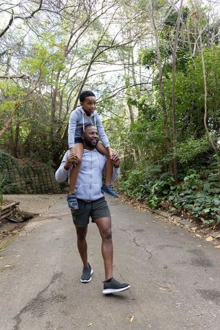 African american father carrying son on shoulders walking along leafy woodland trail