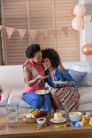 Two friends celebrating birthday on couch sharing cake at cozy living room party