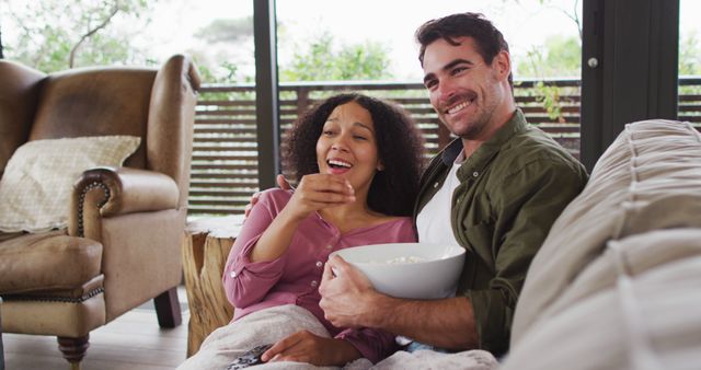 Smiling biracial couple eating popcorn while watching a movie together at vacation home - Download Free Stock Photos Pikwizard.com