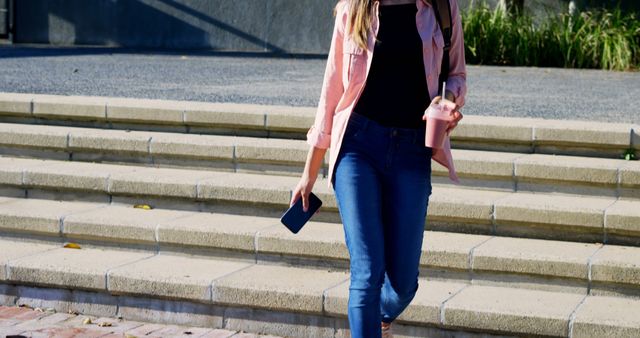 Young woman enjoying a sunny day, walking outdoors with a pink smoothie in one hand and a smartphone in the other. She is dressed in casual wear, including jeans and a light pink shirt. This can be used for lifestyle blogs, advertisements related to beverages or fitness, and any content promoting casual and modern lifestyles.