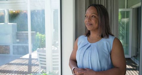 Mature african american woman standing by sliding glass door watching sunlit backyard patio