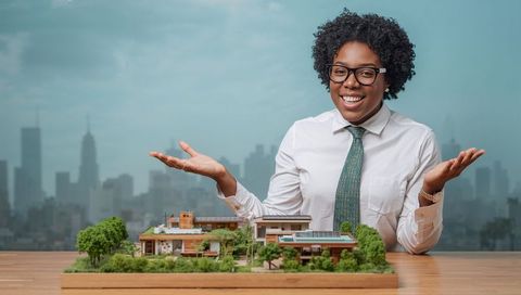 Presenting black architect showcasing sustainable residential scale model with city skyline backdrop