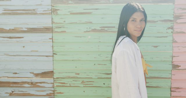 Young woman stands by a colorful, distressed wooden wall, smiling, and wearing a white shirt. The image, with its mix of vibrant pastel colors, gives a lively yet relaxed and casual vibe. Perfect for promotions related to fashion, lifestyle blogs, outdoor activities, urban settings, or marketing materials that aim to convey happiness and relaxation.