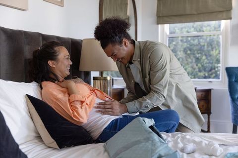 Expectant couple laughing on bed while partner listening to baby bump