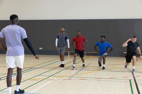Multiracial men doing lunges in indoor gymnasium with coach leading fitness drill