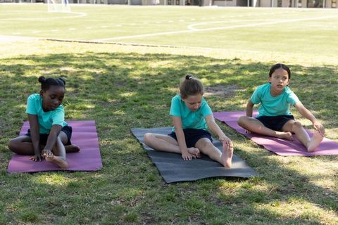 Diverse schoolgirls stretching on mats outdoors during pe class on grassy field