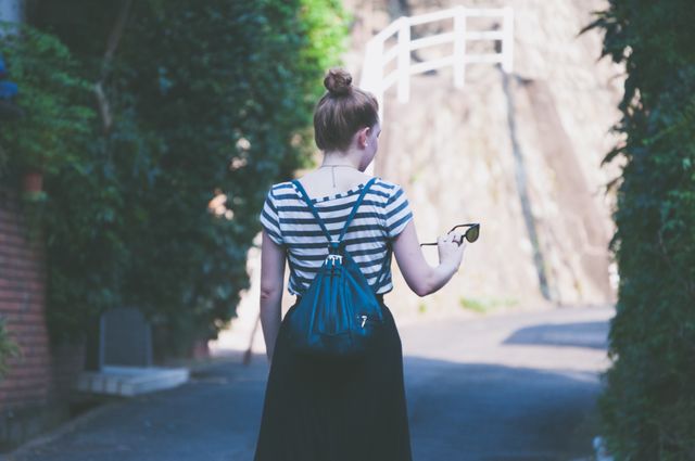 Woman walking down narrow alley holding sunglasses, wearing striped shirt and carrying backpack. Ideal for use in travel blogs, urban exploration themes, adventure promotions, solo travel campaigns, or lifestyle magazines.