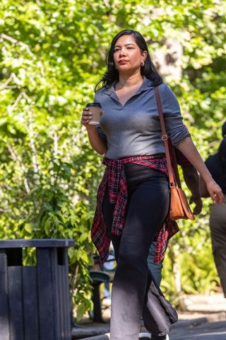 Woman walking through sunlit park holding coffee, wearing red plaid shirt tied at waist