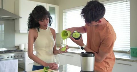 Diverse couple pouring bright green smoothie at modern kitchen island, healthy breakfast