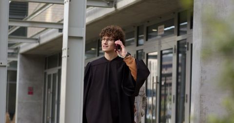 Young graduate walking campus talking on smartphone in academic gown and glasses