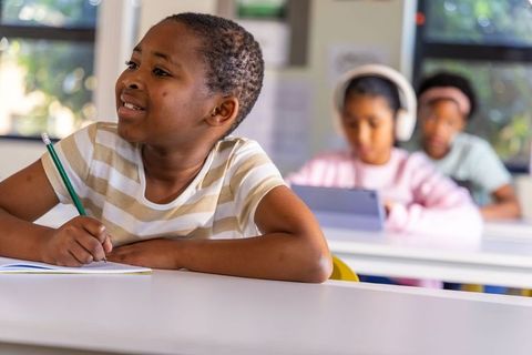Focused african american pupil writing with classmates using tablet and headphones