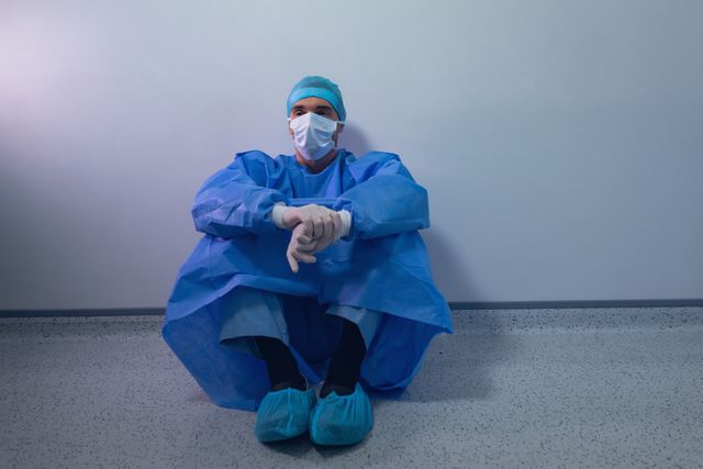 Surgeon in blue scrubs and mask sitting on floor in operating room, appearing thoughtful and contemplative. Useful for illustrating themes of healthcare, medical professionals, stress, and the demanding nature of medical work. Ideal for articles, blogs, and presentations on healthcare challenges, mental health of medical staff, and hospital environments.