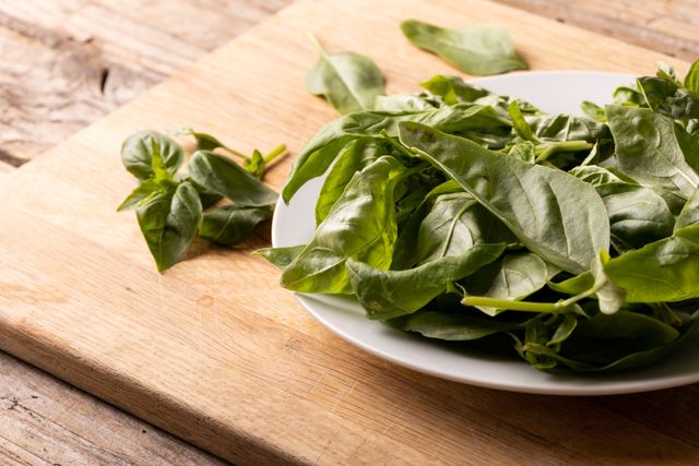 High angle view of fresh spinach leaves on a white plate placed on a wooden cutting board. Ideal for use in articles or blogs about healthy eating, organic food, vegan recipes, or clean eating. Perfect for illustrating concepts related to natural food, cooking ingredients, and healthy lifestyle choices.