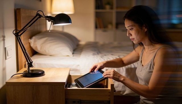 Asian woman retrieving tablet from bedside drawer at night with warm lamp glow and focus