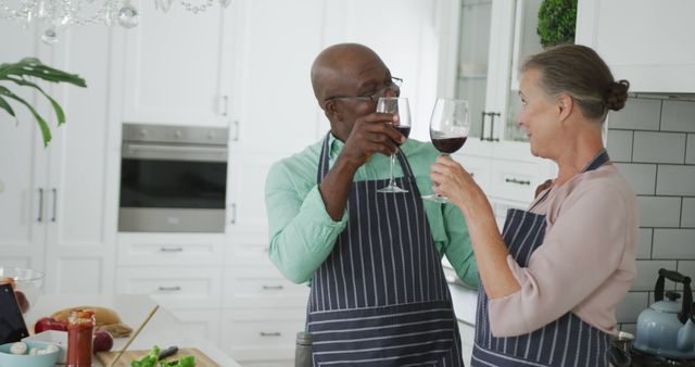 Senior couple wearing aprons and toasting with red wine while cooking together in a modern kitchen. Great for promoting healthy relationships, cooking classes, and home lifestyle products.
