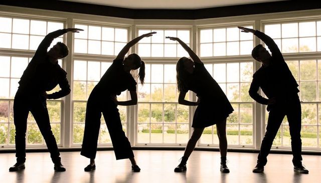 Backlit dance quartet stretching by bay windows, silhouetted rehearsal choreography