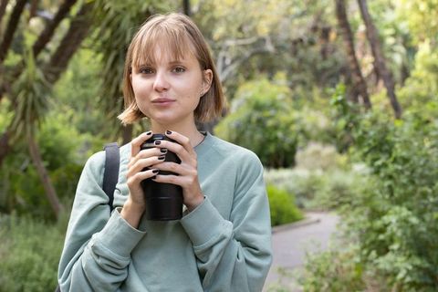 Young woman holding travel mug on park pathway wearing green sweater and backpack smiling softly