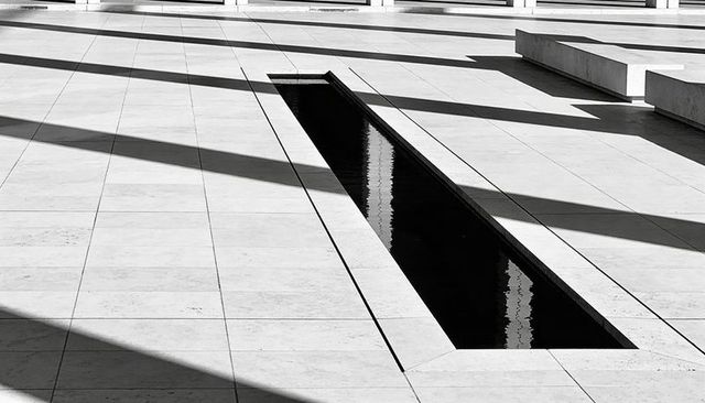 Minimalist geometric reflecting pool with strong diagonal shadows on marble plaza benches