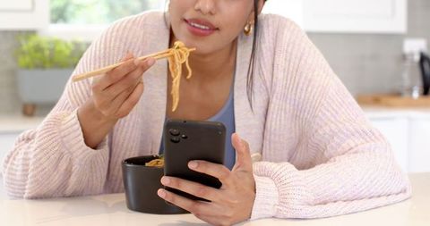 Woman Enjoying Noodles While Using Smartphone in Kitchen