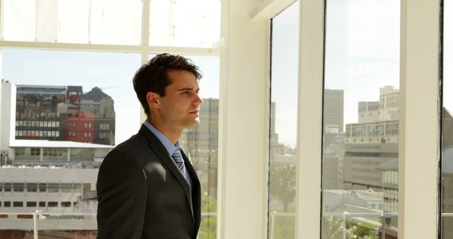 A businessman is standing alone in a modern office with large windows that offer a clear view of the city's skyscrapers. His reflective pose and the natural sunlight streaming through the windows create a bright and contemplative atmosphere. This visual can be used for business presentations, articles about corporate life, leadership, decision-making, or personal growth within a professional context.