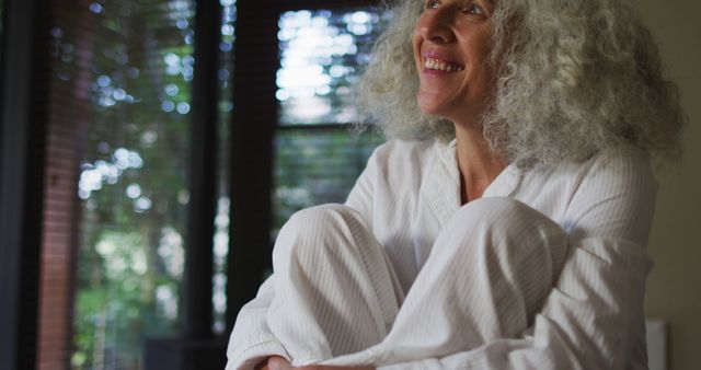 Senior woman with curly gray hair smiling while sitting by window wearing pajamas. Natural light flooding the room, creating a serene atmosphere. Ideal for use in health and wellness articles, aging gracefully topics, senior lifestyle promotions, and content on happiness and relaxation.