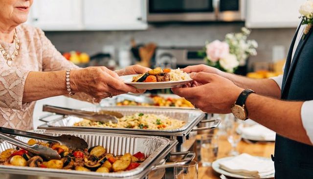 Elderly woman handing plate to man in vest at elegant home buffet during family celebration