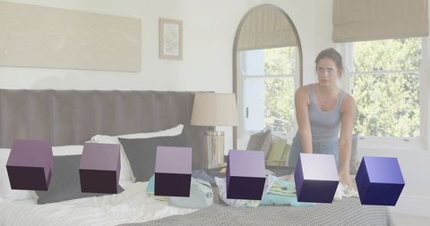 Young woman leaning over bed packing suitcase in sunlit modern bedroom with purple cubes