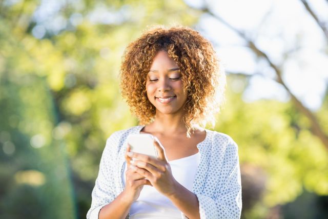 Woman standing in a sunny park, smiling while using her mobile phone. Ideal for themes related to technology, communication, outdoor activities, and lifestyle. Perfect for advertisements, blog posts, and social media content focusing on mobile technology, leisure activities, and positive emotions.