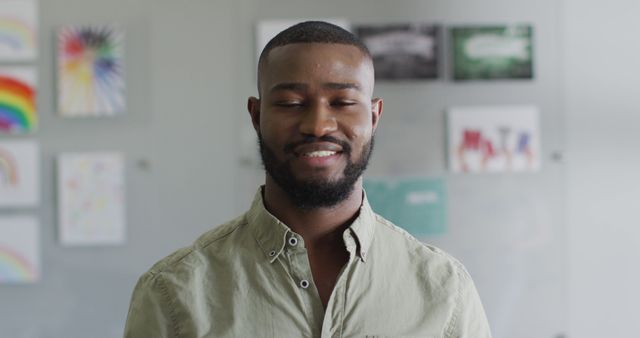 Close-up of a smiling young man in a casual shirt standing indoors with various artworks displayed on the wall behind him. Perfect for use in lifestyle blogs, home decor advertisements, or online articles about personal happiness and relaxation.
