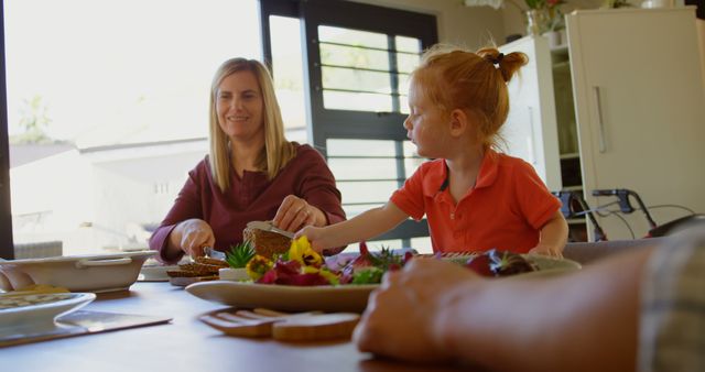Image shows mother and daughter enjoying lunch together at home. The scene captures a warm family moment during a casual dining setting brightened by natural light. Suitable for use in family-focused content, parenting articles, meal time routines, family vlogs, and advertisements promoting home-cooked meals or family bonding time.