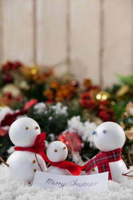Three snowmen with red and plaid scarves are surrounded by fake snow and festive decorations. A card with 'Merry Christmas' written on it is placed in front. Ideal for holiday greeting cards, Christmas decorations, and festive marketing materials.