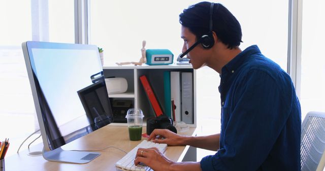 A young Asian man is working at his desk with a headset on, engaged in a customer service or remote communication job, with copy space. His focused demeanor and organized workspace suggest a professional environment.