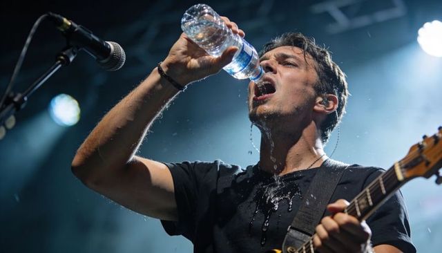 Male musician drinking water onstage while playing guitar under concert lights and mist