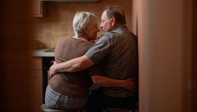Senior couple embracing on kitchen stools sharing intimate cozy domestic moment with warm lighting