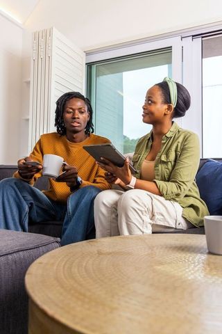 African American Couple Relaxing on Sofa Using Tablet and Drinking Coffee in Bright Apartment