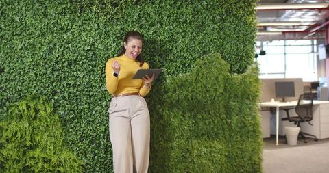 Professional celebrating while using tablet and leaning on green wall in coworking office