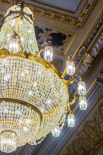 Close-up of luxurious crystal chandelier with gold embellishments hanging in elegant ballroom setting. Ideal for interior design inspiration, luxury event decorators, architectural references, and historical preservation projects.