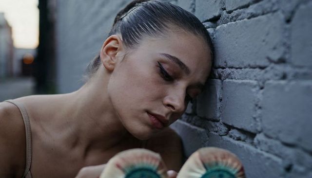 Leaning urban ballerina holding pointe shoes against textured brick wall at dusk